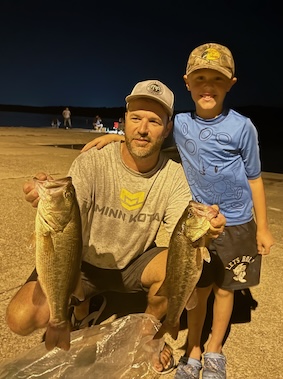 Matt crouching holding two large fish with his young son's hand around his shoulders in a nightime setting near a lake.