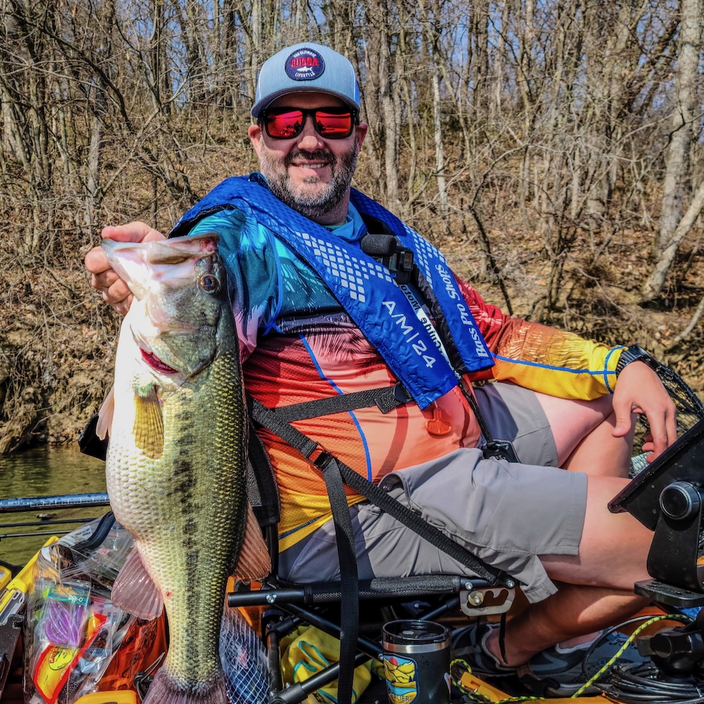 A man on a boat with a bare tree shoreline in the background holding up a large fish.